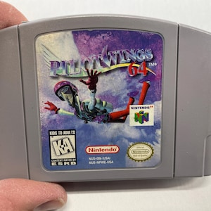 Puede incluir: Un cartucho de juego gris de Nintendo 64 con el juego "Pilotwings 64". La etiqueta muestra un paracaidista en el aire con el título del juego. El logotipo de Nintendo y la clasificación ESRB también son visibles.
