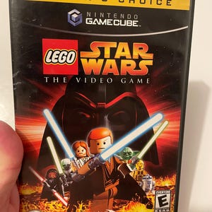 May include: A video game case for the Nintendo GameCube featuring Lego Star Wars: The Video Game. The case is black with a yellow border and features a picture of Lego characters from the Star Wars franchise, including Darth Vader, Yoda, and Obi-Wan Kenobi. The game is rated E for Everyone by the ESRB.