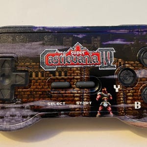 Könnte beinhalten: Ein schwarzer und grauer Super Castlevania IV Videospiel-Controller. Der Controller hat ein Backsteinmauer-Design mit dem Logo des Spiels und Charaktergrafiken. Er hat ein Steuerkreuz, Tasten und den Text "SELECT" und "START".