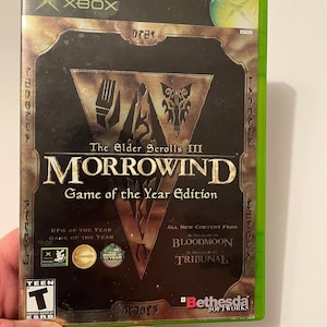 May include: Xbox game case for The Elder Scrolls III: Morrowind Game of the Year Edition. The case is green and features a black and gold design with the game title and logo. The case also includes the ESRB Teen rating and the Bethesda Softworks logo.