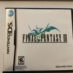 Op de afbeelding: Een witte Nintendo DS-gamecase met een zwarte rand. De game is Final Fantasy III. De gamecase heeft een classificatie van "Voor iedereen 10+" door de ESRB. De gamecase heeft een blauw Nintendo Wi-Fi Connection-logo. De gamecase heeft de tekst "Game Experience May Change During Online Play". De gamecase heeft de tekst "SQUARE ENIX."