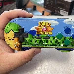 May include: A handheld video game controller with a colorful design featuring the text "Kirby Super Star." The controller has a blue top with blue and green buttons, a black directional pad, and a landscape graphic.