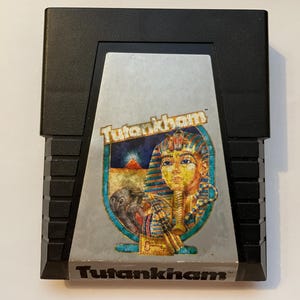 Puede incluir: Cartucho de videojuego negro con una etiqueta plateada que presenta el título del juego "Tutankham" y una ilustración de un faraón. El cartucho tiene una carcasa de plástico negro.