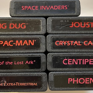Könnte beinhalten: Eine Sammlung schwarzer Atari-Spielmodule mit roter Schrift. Zu den Titeln gehören "Space Invaders", "Dig Dug", "Ms. Pac-Man", "Crystal Castles", "Raiders of the Lost Ark", "Centipede", "E.T. The Extra-Terrestrial", "Phoenix" und "Joust".