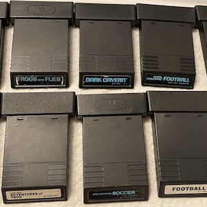 May include: A collection of ten black Nintendo Entertainment System (NES) game cartridges. The cartridges are arranged in two rows of five. The game titles are: Astroblast, Frogs and Flies, Dark Cavern, Football, Baseball, Space Attack, Adventures of Tron, Soccer, Football, and Lock 'n' Chase.