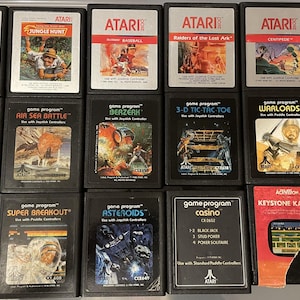 Può includere: Una collezione di 14 cartucce di videogiochi Atari vintage, tra cui titoli come Jungle Hunt, Raiders of the Lost Ark e Keystone Kapers. Le cartucce sono disposte in tre file di quattro, con uno sfondo nero.