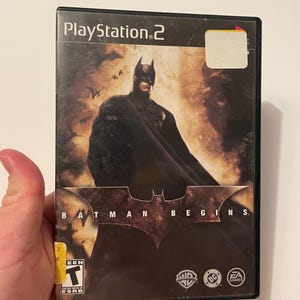 Puede incluir: Una caja de juego de PlayStation 2 negra con una imagen de fondo oscura de Batman con una capa. El título del juego es "Batman Begins" y el juego está clasificado como "T" para adolescentes por la ESRB. La caja también presenta los logotipos de Warner Bros., DC Comics y EA Games.