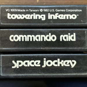 May include: Three vintage video game cartridges stacked on top of each other. The cartridges are black with white text. The top cartridge reads "towering inferno", the middle cartridge reads "commando raid", and the bottom cartridge reads "space jockey".