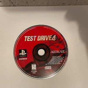Op de afbeelding: Een rode PlayStation-gamedisc voor Test Drive 4, een racespel uitgegeven door Acclaim Entertainment. De disc heeft een zilveren midden met een zwarte cirkel in het midden. De disc is gelabeld met de gametitel, uitgever en beoordeling.