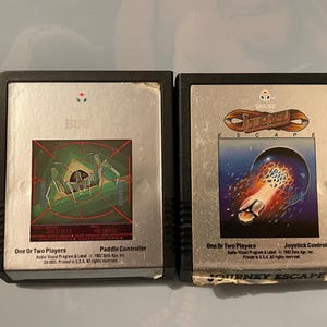 May include: Two Atari 2600 game cartridges, one for Journey Escape and one for Bug. Both cartridges have black plastic cases with silver labels. The Journey Escape cartridge has a blue and white label with a graphic of a spaceship flying through space. The Bug cartridge has a green and white label with a graphic of a bug.