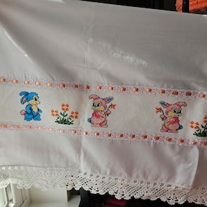 May include: White fabric with a pink and white lace trim. A row of embroidered animals with flowers is stitched along the top edge. The animals are blue, pink, and green.