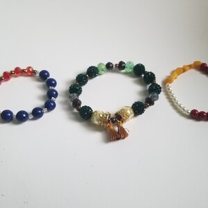 Puede incluir: Tres pulseras de cuentas con cuentas de diferentes colores. La primera pulsera tiene cuentas rojas y azules. La segunda pulsera tiene cuentas verdes y doradas con un borla dorada. La tercera pulsera tiene cuentas ámbar, blancas y rojas.