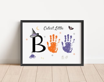 Puede incluir: Un estampado enmarcado con temática de Halloween con las palabras "Cutest Little Boo" y arte de huellas de manos. La letra "B" es grande y negra, con huellas de manos naranjas y moradas. Se incluye un sombrero de bruja, luna, estrellas y otros elementos de Halloween.