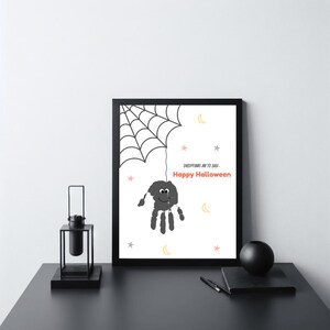 Pode incluir: Uma impressão emoldurada com tema de Halloween com uma aranha de impressão de mão pendurada em uma teia. A impressão inclui o texto "Happy Halloween" em vermelho, com estrelas e luas. A moldura é preta.
