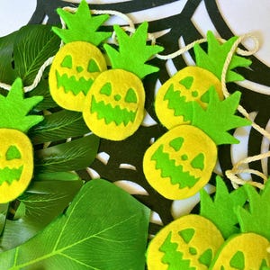 May include: A string of felt pineapple decorations with jack-o'-lantern faces. The pineapples are yellow with green leaves and green faces. The pineapples are strung together with white twine.