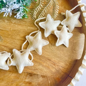 May include: A string of off-white felt stars on natural twine. The stars are arranged in a row, with a rustic wooden tray in the background. Decorative greenery is visible in the upper left corner. The garland is a festive decoration.