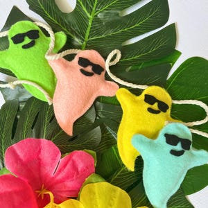 May include: Four felt ghost decorations with sunglasses, each in a different colour: green, pink, yellow, and blue. The ghosts are hanging from a white string and are displayed against a green tropical leaf background.