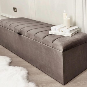 May include: A grey velvet storage bench with a tufted top and a hinged lid. The bench is sitting on a white rug and has books and candles on top.