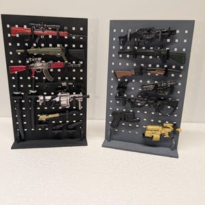 May include: Two display stands showcasing miniature toy guns. One stand is black, the other is gray. The toy guns are various colors, including red, green, black, and gold. The stands are on a white surface.