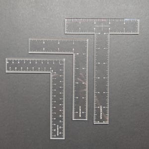 May include: Four clear acrylic T-squares and rulers of varying sizes are arranged on a gray surface. The rulers have white markings for inches and fractions of an inch. The largest T-square has a 12-inch vertical ruler.