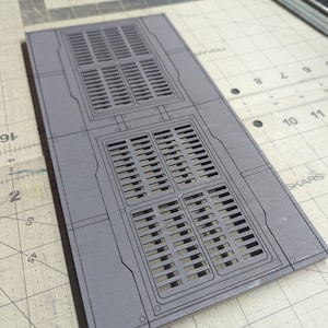May include: A rectangular, gray panel with two sets of rectangular vents. The panel has a series of etched lines and small circular details. The design suggests a futuristic or industrial theme.