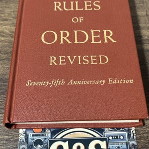 May include: A hardcover book titled Robert's Rules of Order Revised, Seventy-fifth Anniversary Edition, with a textured brown cover and gold lettering. Below the book is a rectangular card featuring the text G&S VINTAGE MEDIA.