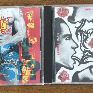 May include: Two CD cases. The first features the text "Red Hot Chili Peppers" and "What Hits?!" on a red background with a collage of faces. The second CD case has a black and white design with faces and roses, with the text "Red Blood" and "Hot Sugar".