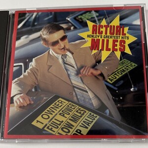 May include: CD case featuring the album "Actual Miles: Henley's Greatest Hits." The cover art shows a man in a tan suit and sunglasses leaning on a car. The album title is in a yellow starburst.