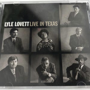 May include: A black and white CD case featuring the text "LYLE LOVETT LIVE IN TEXAS". The cover displays a grid of six black and white portraits of people. The CD case is in a clear plastic jewel case.