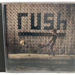 May include: A Rush Roll the Bones music CD in a clear plastic jewel case. The cover art features a person kicking a floating skull in front of a wall constructed from white dice. The band name RUSH and the album title are printed on the wall.