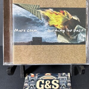 May include: A Marc Cohn Burning The Daze music CD in a clear plastic jewel case. The cover art features a paper airplane on fire against a dark sky and a silhouette of a house. Below the CD is a rectangular card with the text G&S VINTAGE MEDIA.