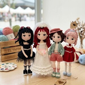 May include: Four handmade crochet dolls with detailed outfits. One doll wears a black dress, another a white wedding dress, a third doll is in a green jacket and shorts, and the last doll is in a red dress. The dolls have yarn hair and are standing on a wooden surface.