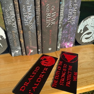 May include: A selection of fantasy novels with matching black bookmarks. The books are titled "Of Empires and Dust" and "Of Blood and Fire." The bookmarks have red text and dragon or triangle designs, with phrases like "The Duty of the Strong."