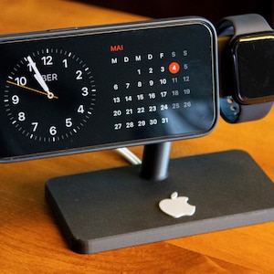 May include: A black charging stand with a white Apple logo, holding a black iPhone with a clock face and calendar showing the month of May, and a black Apple Watch.