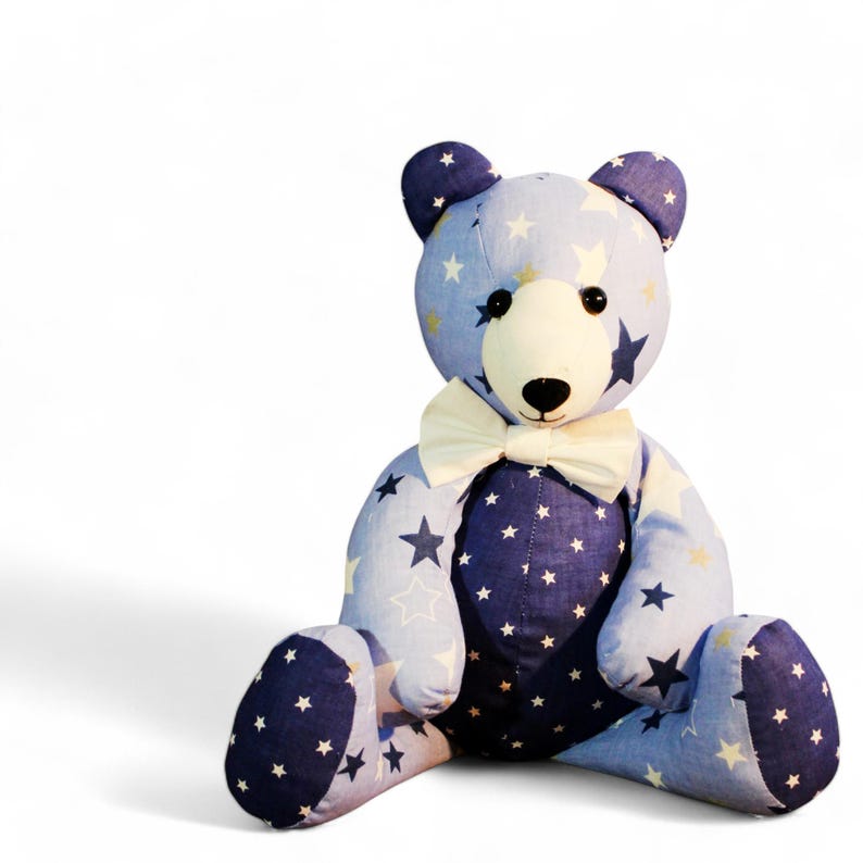 May include: A handmade teddy bear with a light blue body and dark blue accents, adorned with white and yellow stars. The bear has a cream-colored face and a white bow tie. A charming and whimsical plush toy.
