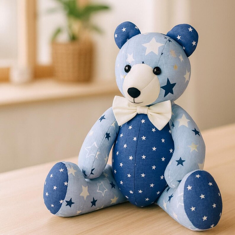 May include: A plush teddy bear with a light blue body and dark blue accents, adorned with white stars. The bear has a white bow tie and black button eyes, sitting on a wooden surface. A charming, handmade children's toy.