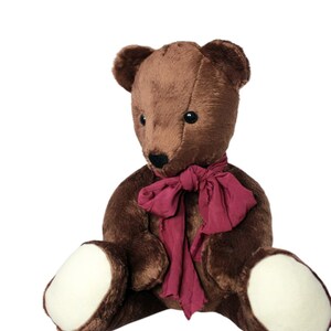 May include: A brown teddy bear with a dark red scarf. The bear has a black nose and eyes, and white patches on its feet. The plush toy is sitting down and has a classic, vintage appearance.