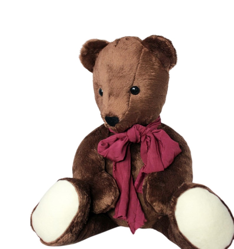 May include: A brown teddy bear with a dark red scarf. The bear has a black nose and eyes, and white patches on its feet. The plush toy is sitting down and has a classic, vintage appearance.
