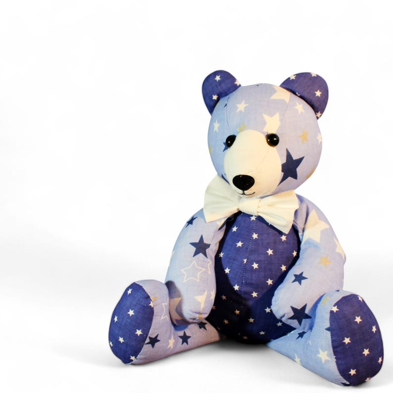 May include: A plush teddy bear with a light blue and navy blue star pattern. The bear has a white bow tie and a cream-colored snout. The bear is sitting with its legs extended and arms at its sides.
