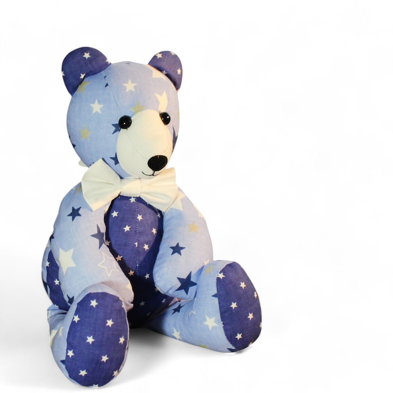 May include: A plush teddy bear with a light blue body and dark blue accents, patterned with white and yellow stars. The bear has a cream-colored face, black eyes, and a white bow tie. It is sitting upright.