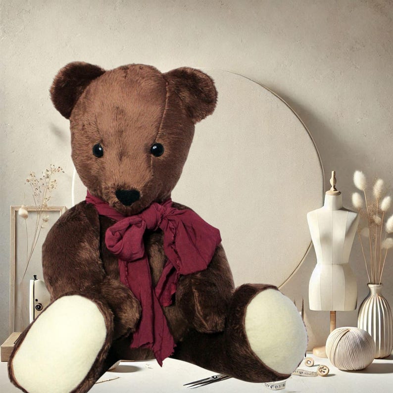 May include: A large, brown teddy bear wearing a dark red scarf. The bear has a soft, plush appearance with light-coloured paws. The background includes a mannequin, a vase with dried flowers, and a ball of yarn, suggesting a craft or sewing theme.