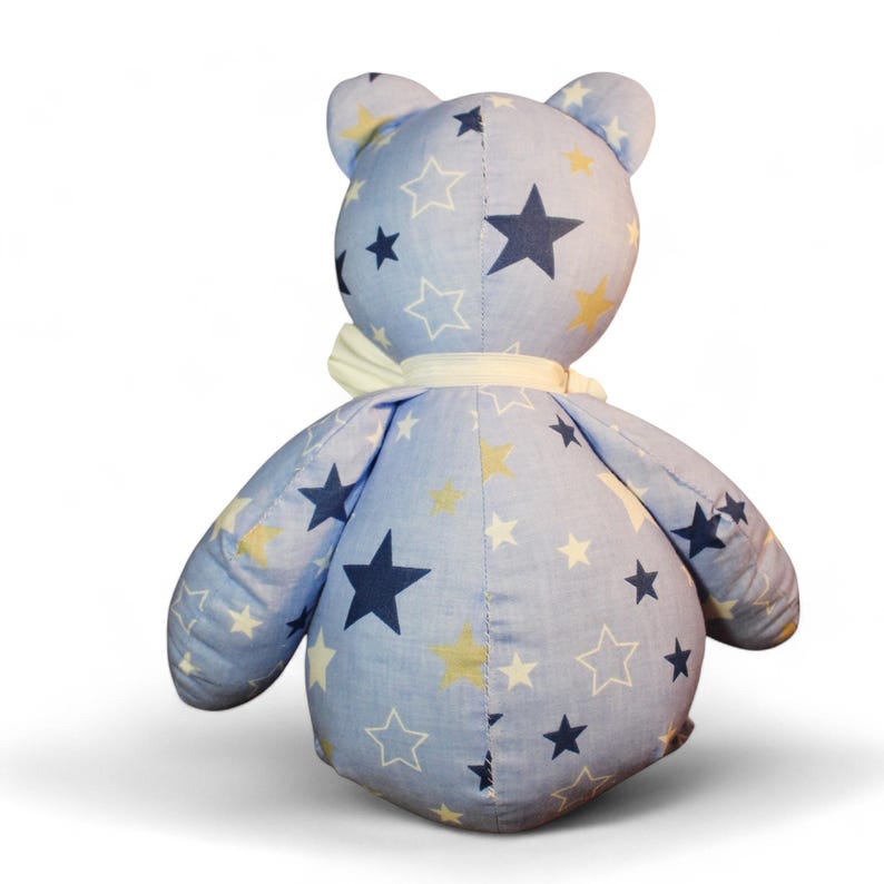May include: A handmade stuffed bear with a light blue fabric covered in white, navy, and yellow stars. The bear has a white ribbon around its neck. The bear is a soft toy.