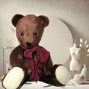 May include: A large brown teddy bear wearing a dark red scarf. The bear has a black nose and cream-colored paws. The background features a dress form, a vase with dried flowers, and a ball of yarn, all in a neutral color palette.