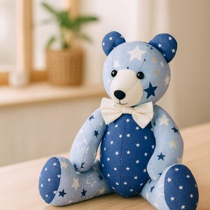 May include: A plush teddy bear with a light blue and navy blue star pattern. The bear has a white muzzle and a cream-colored bow tie. The bear is sitting on a wooden surface.