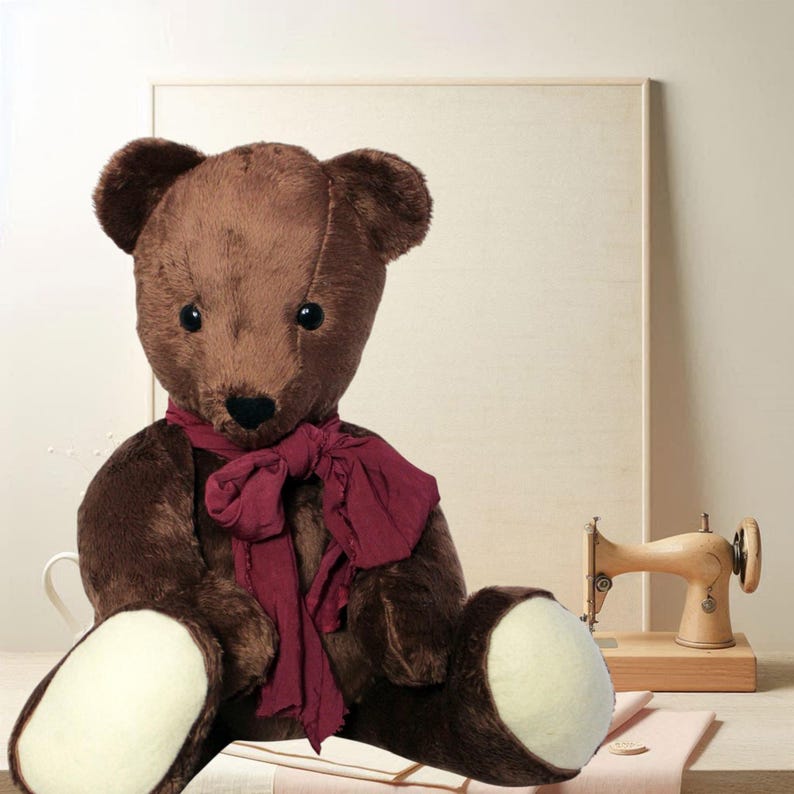May include: A brown teddy bear with a burgundy scarf, sitting on a table. The bear has a stitched nose and button eyes. A vintage wooden sewing machine and a framed canvas are in the background. The bear's paws are cream-coloured.