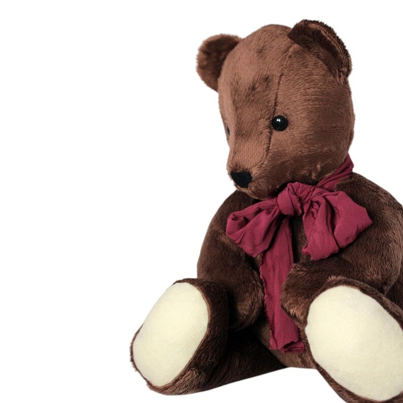 May include: A brown teddy bear wearing a burgundy scarf. The bear has black eyes and cream-coloured paws. The soft toy is set against a white background.