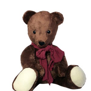 May include: A brown teddy bear wearing a dark red scarf. The soft toy has a black nose, dark eyes, and cream-coloured foot pads. The bear is sitting upright against a white background.