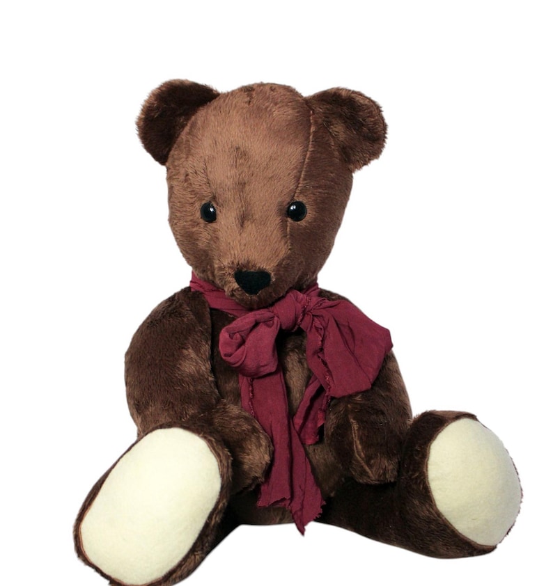 May include: A brown teddy bear wearing a dark red scarf. The soft toy has a black nose, dark eyes, and cream-coloured foot pads. The bear is sitting upright against a white background.