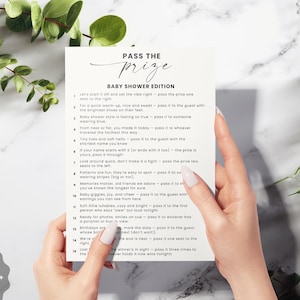 May include: A white baby shower game card titled "Pass the Prize" with a list of instructions, held by a person's hands. The card is surrounded by green leaves and sits on a marble surface. The design includes a logo that says "Happily Ever Design Studio."