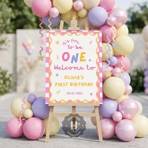 Könnte beinhalten: Eine Holzstaffelei zeigt ein Schild mit dem Text "It's fun to be ONE" für einen ersten Geburtstag. Das Schild ist von einem farbenfrohen Ballonbogen in Rosa-, Gelb- und Lilatönen umgeben. Das Datum auf dem Schild ist der 03-03-20XX.
